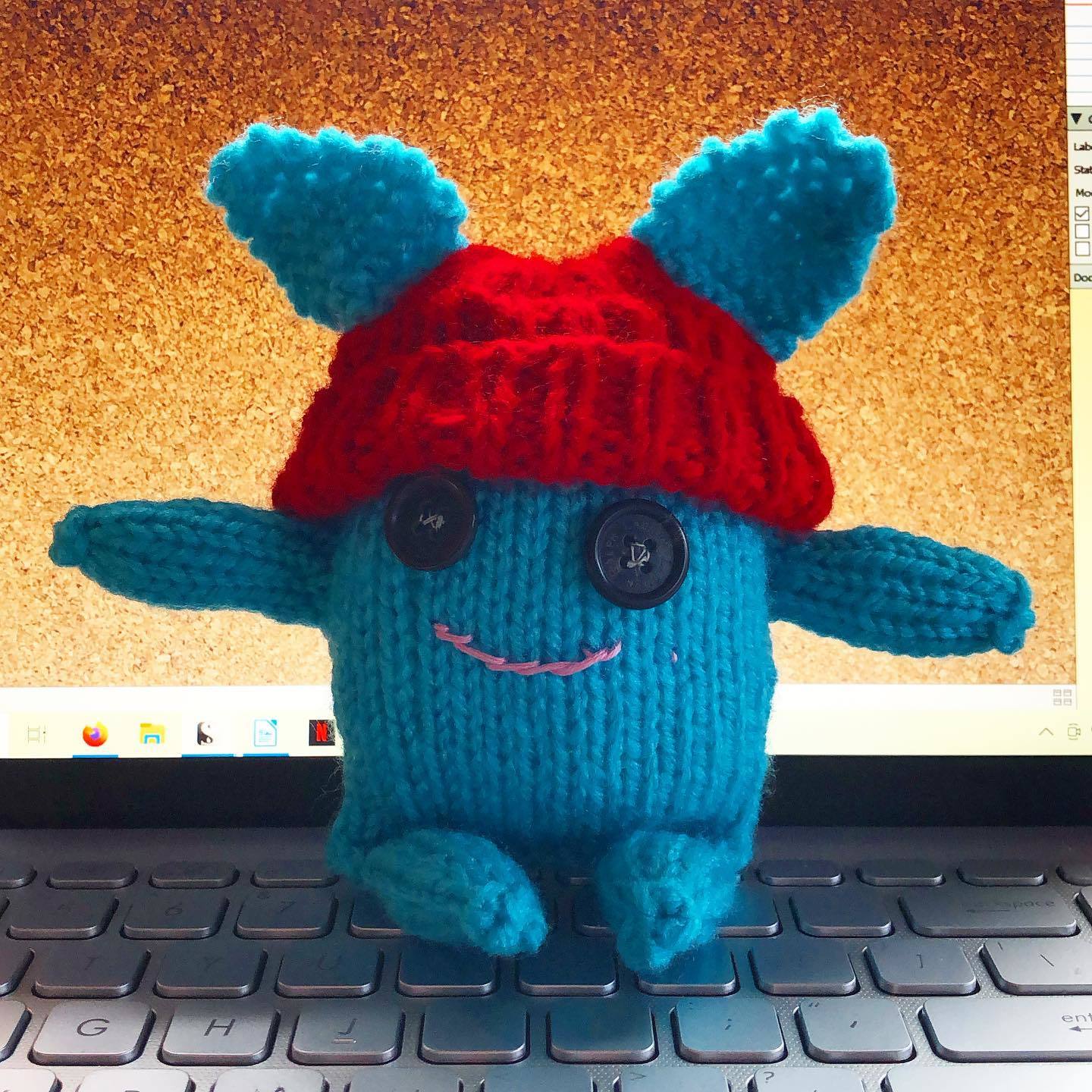 a small knitted dude in blue yarn with a red hat and button eyes